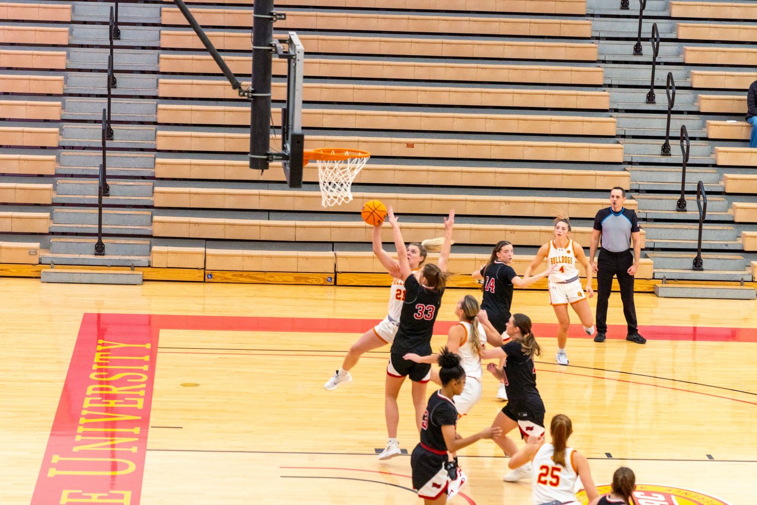 High school basketball players compete in a gymnasium with orange bleachers and spectators watching from the sidelines.
