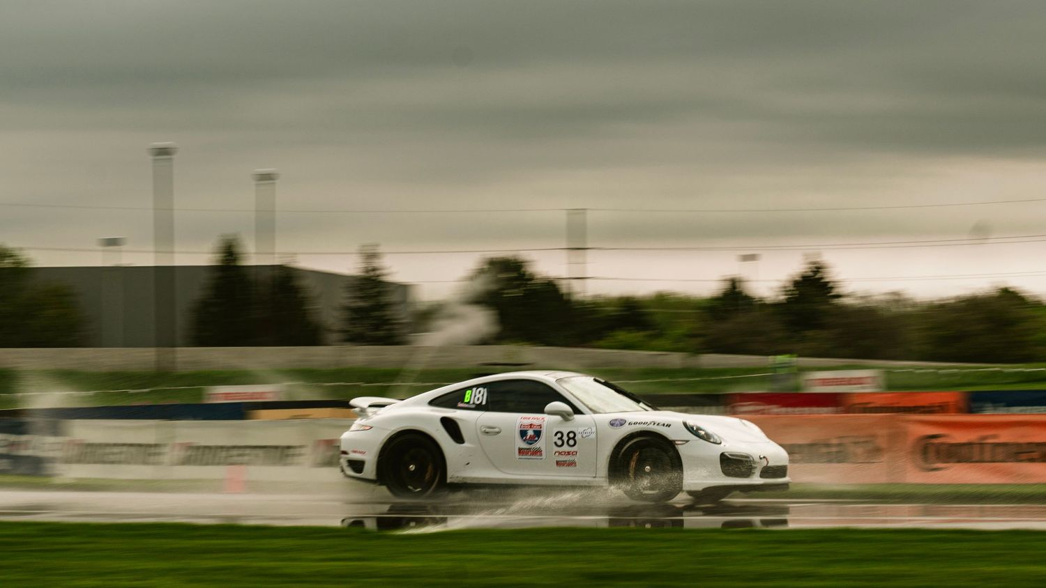 White sports car racing past orange barriers in wet conditions.