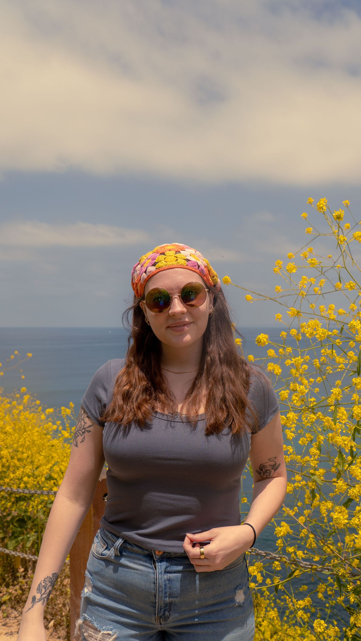 Person in gray shirt and patterned headscarf stands among coastal wildflowers with ocean behind.