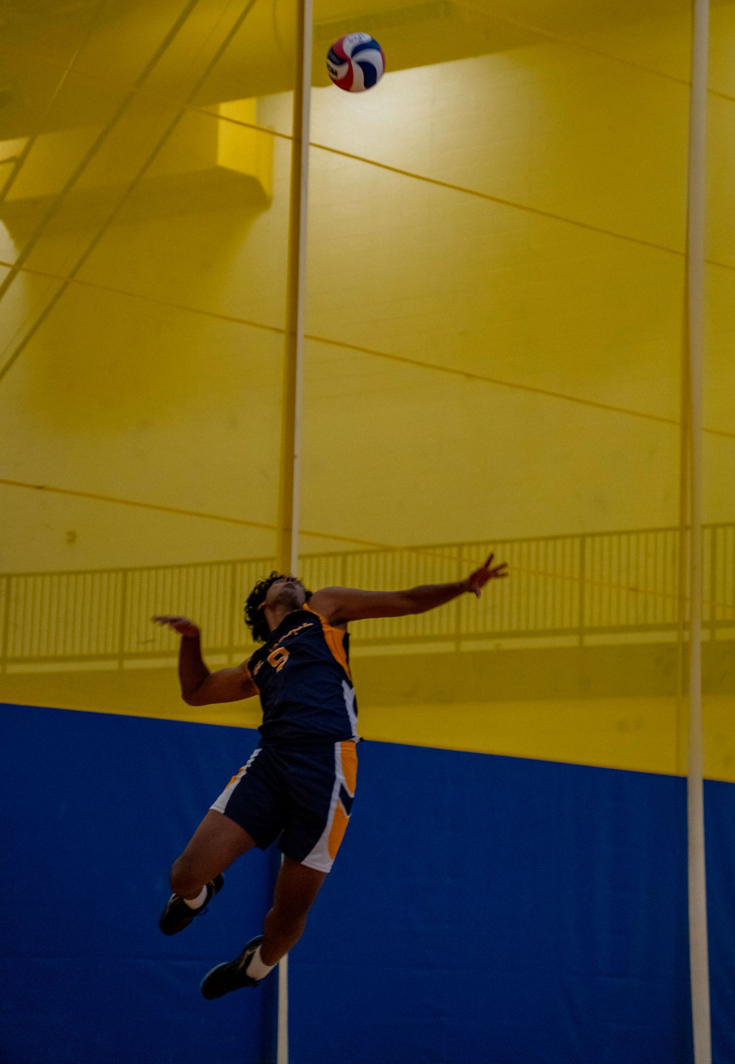 Volleyball player in navy uniform jumping high to spike the ball against yellow gymnasium backdrop.