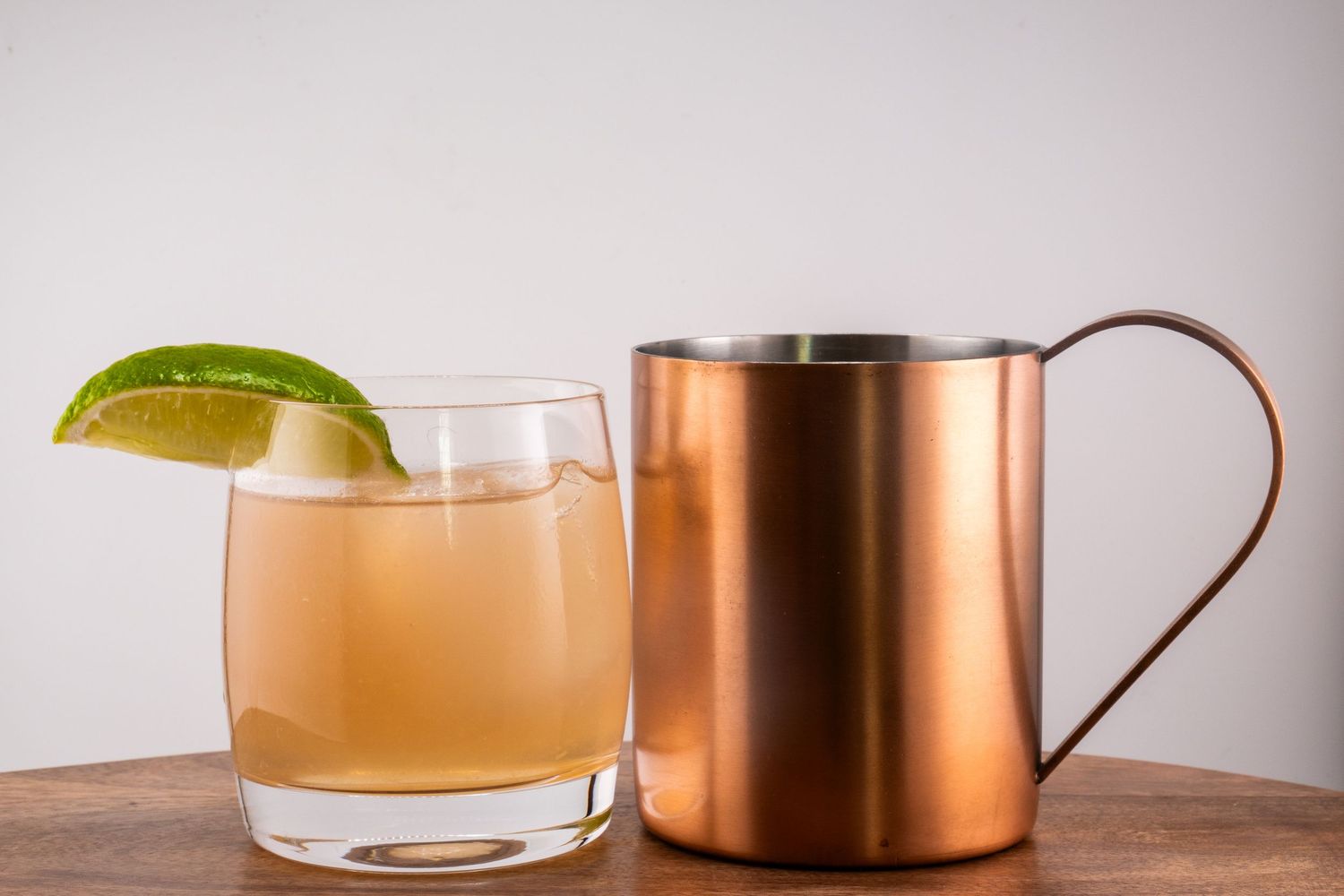 A cocktail glass with lime garnish next to a copper Moscow mule mug on a wooden surface.
