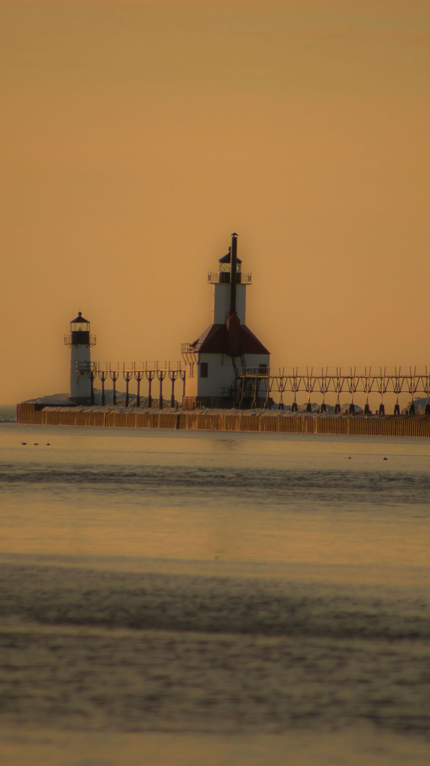 Scenic lighthouse with catwalk pier silhouetted against an orange sunset sky reflecting on calm waters.