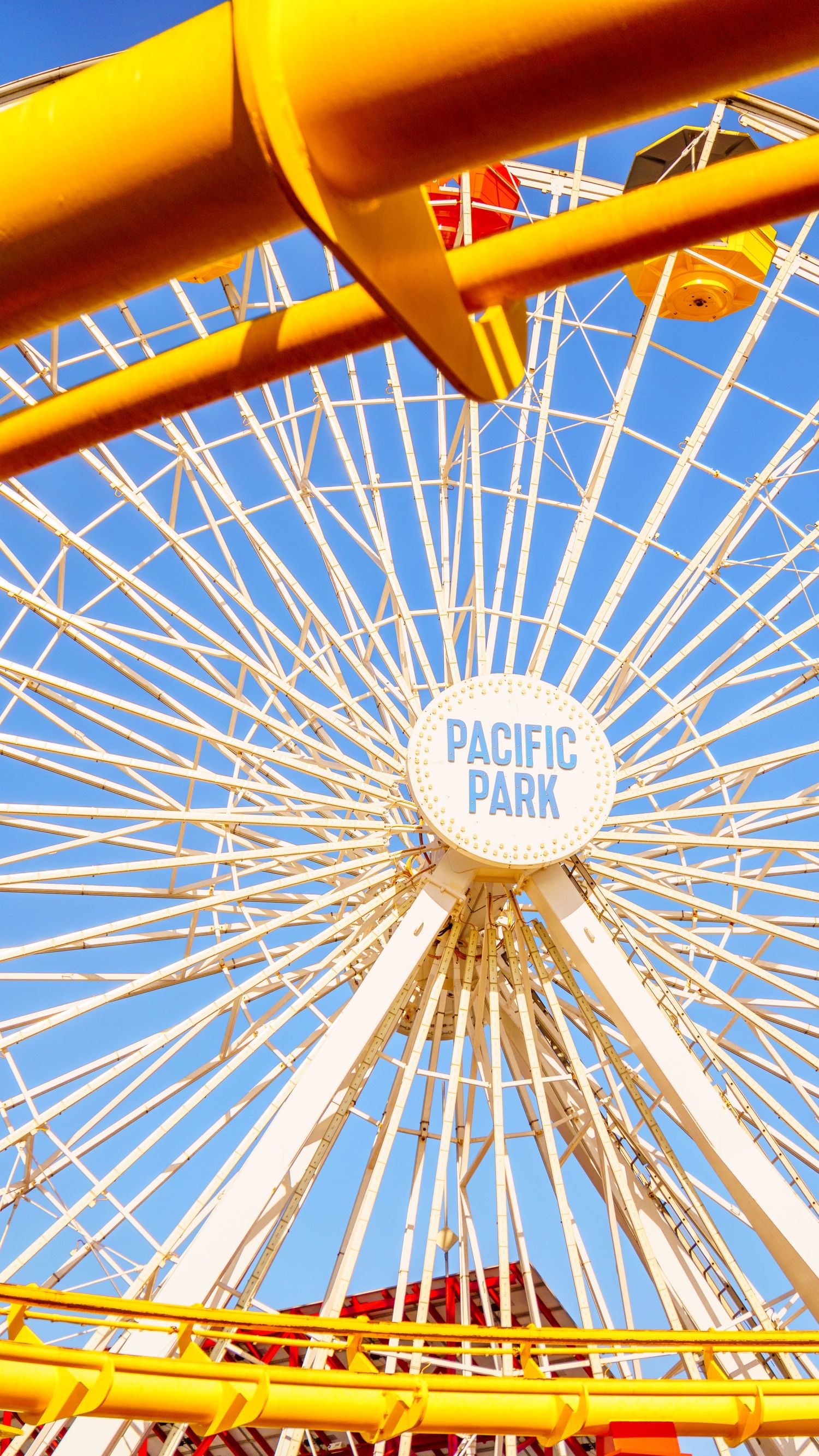 A Pacific Park Ferris wheel's white spokes radiate against a bright blue sky with yellow support beams.