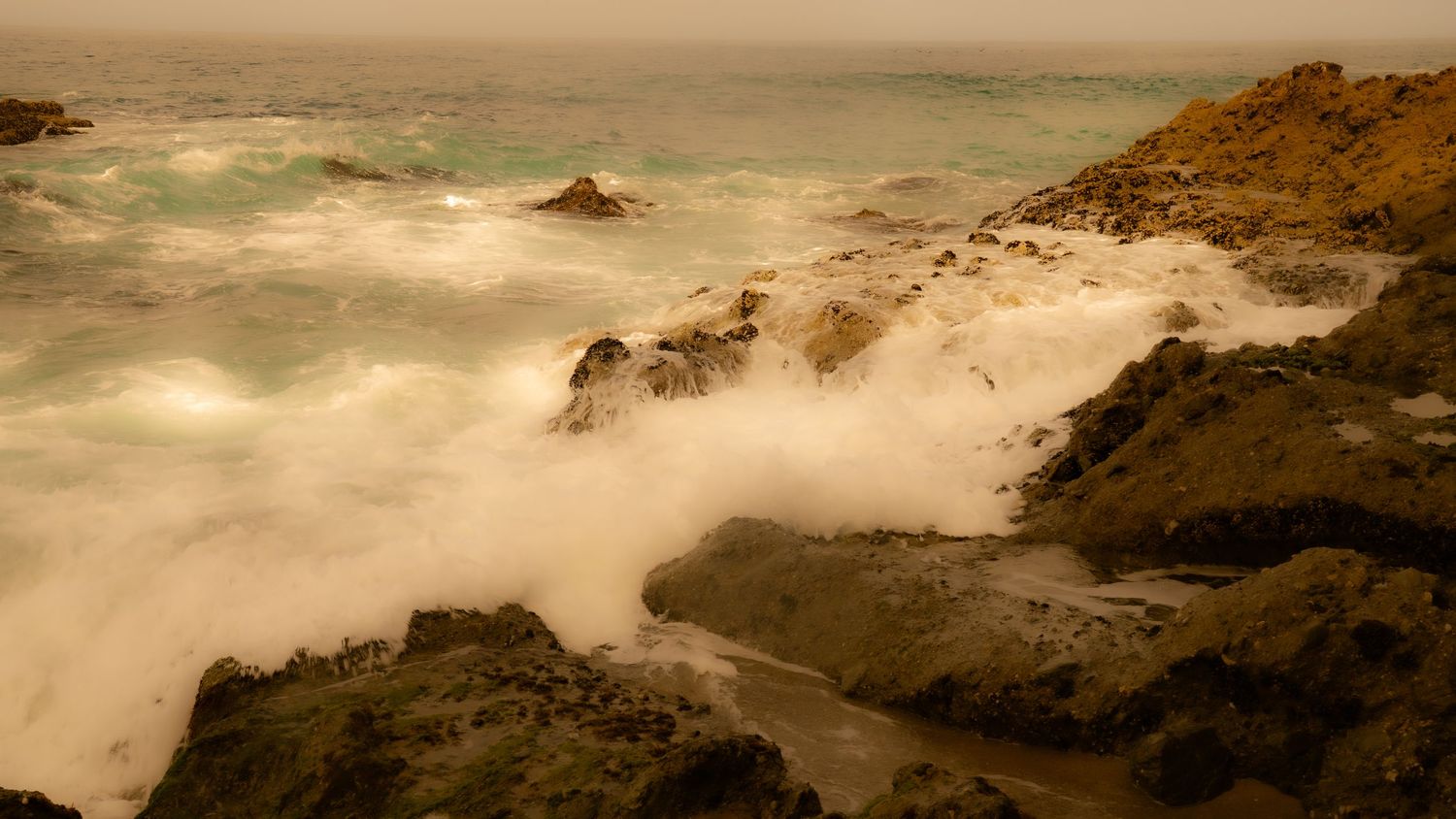 Sepia-toned seascape showing powerful waves crashing against rocky coastal cliffs in misty conditions.