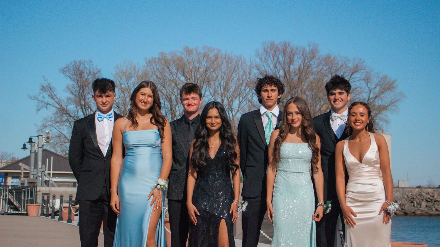 A group in formal attire poses together by the water on a sunny day.