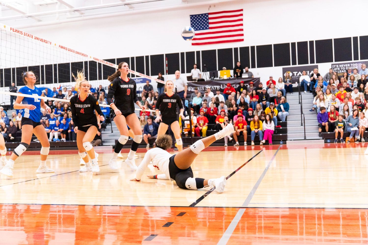 A volleyball player in white dives to the floor for a save as teammates in black and an opponent in blue look on in a packed gymnasium.