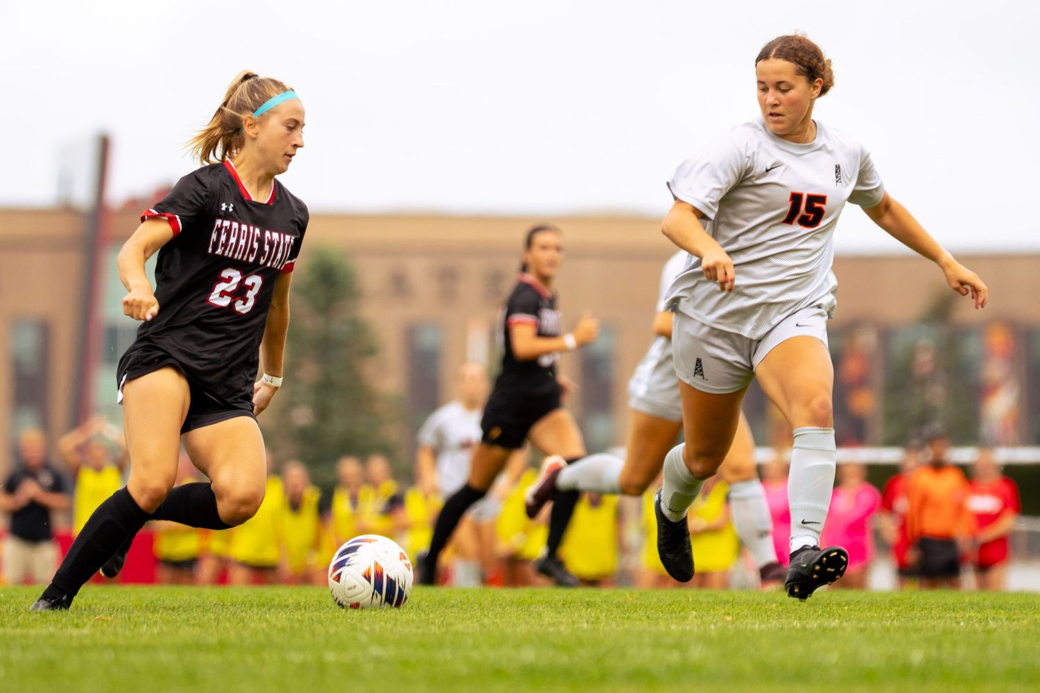 Two women's soccer players chase the ball on a grass field, one in a black number 23 jersey and one in a gray number 15 uniform.
