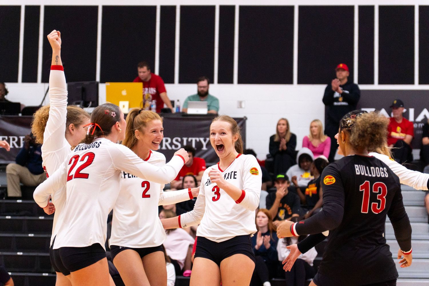 Women's volleyball players in white jerseys celebrate a point on the court while a Bulldogs player in black stands nearby and fans watch.