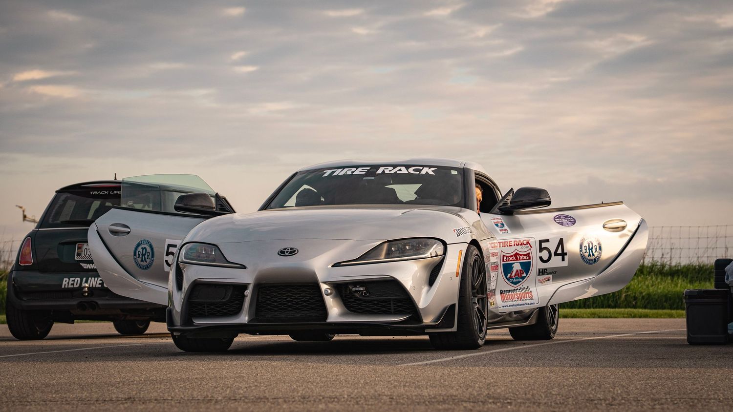 White Toyota Supra race car number 54 parked with other vehicles at sunset.