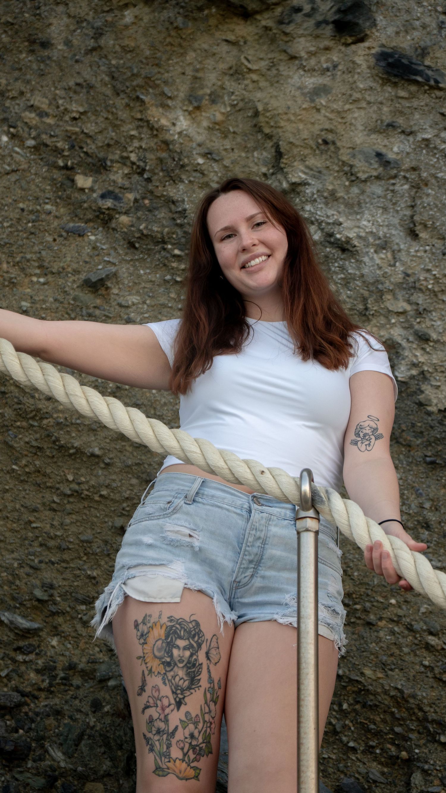 Person in white top and denim shorts posing with rope railing against stone wall.