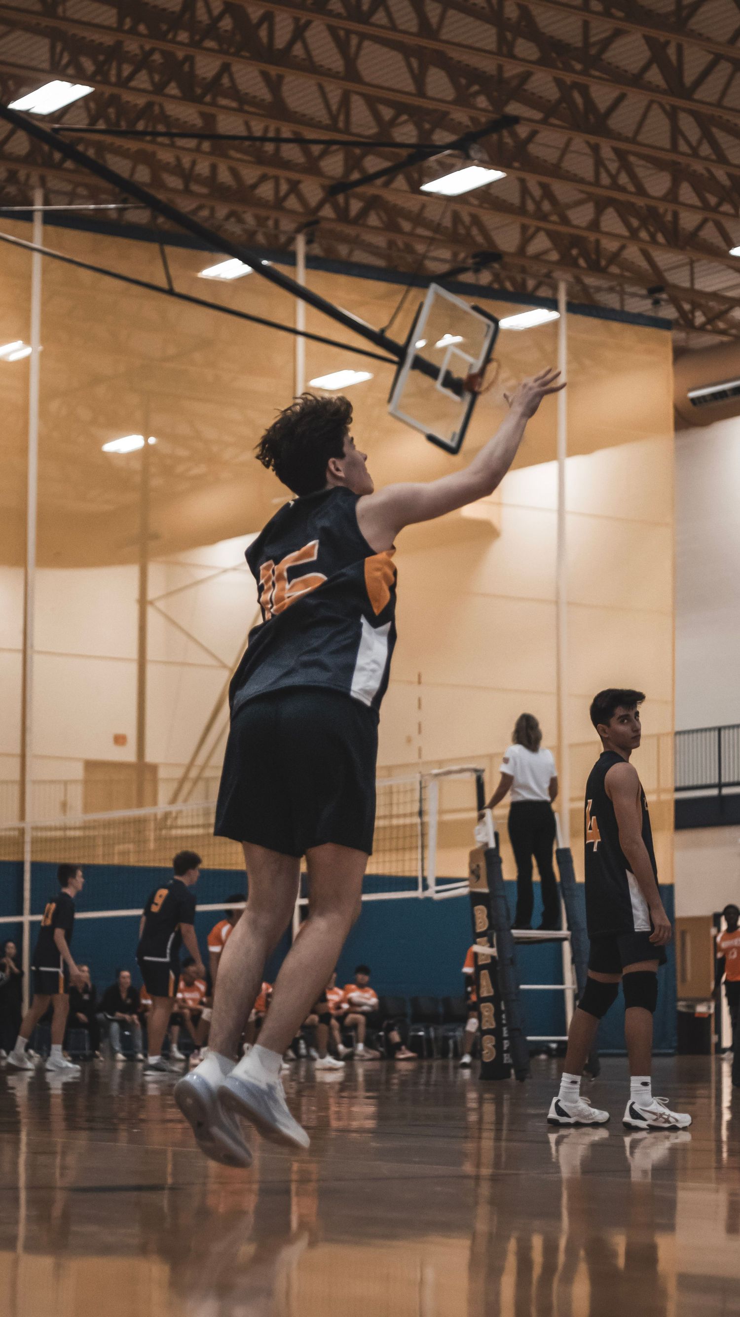 A player in black uniform jumps to serve the volleyball in an indoor gymnasium.