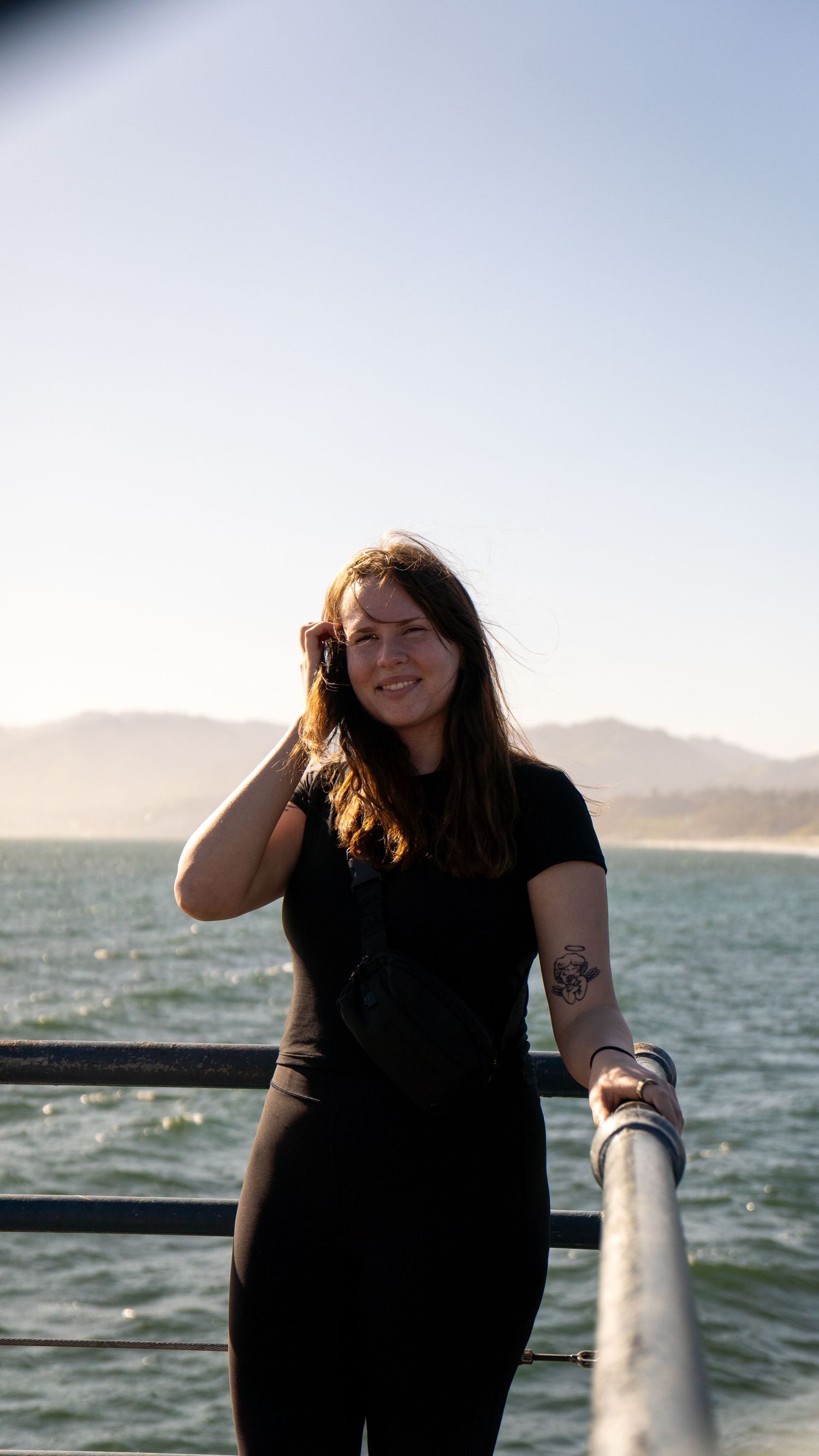 Person in black outfit poses against ocean backdrop during golden hour.