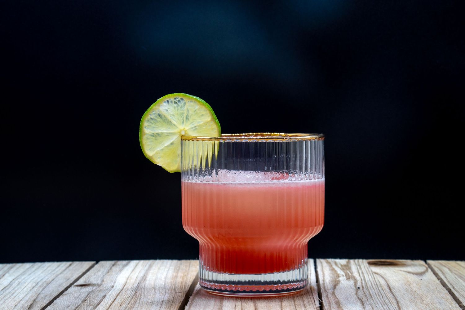 Pink cocktail in a textured glass with lime garnish against dark background.
