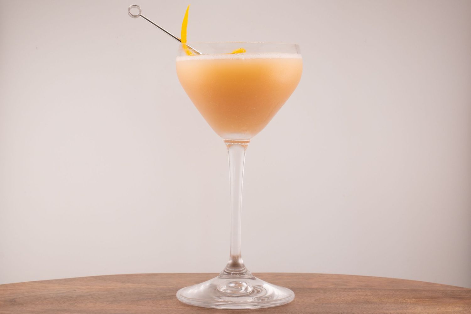 Pastel orange cocktail served in coupe glass with citrus garnish and metal pick.
