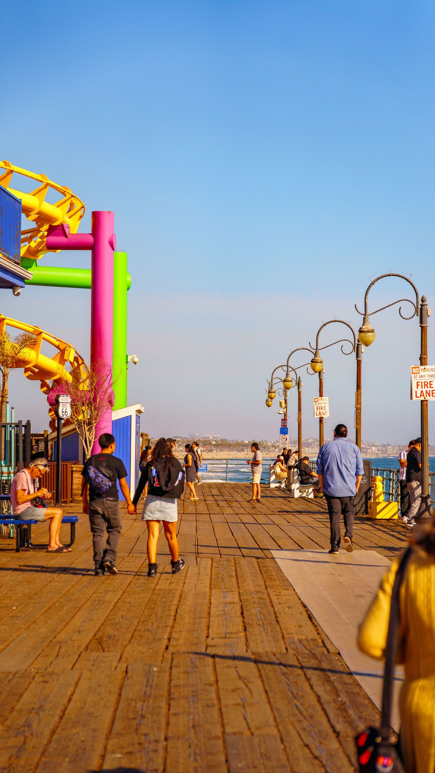 Wooden pier walkway with vintage lampposts and colorful amusement rides in background.