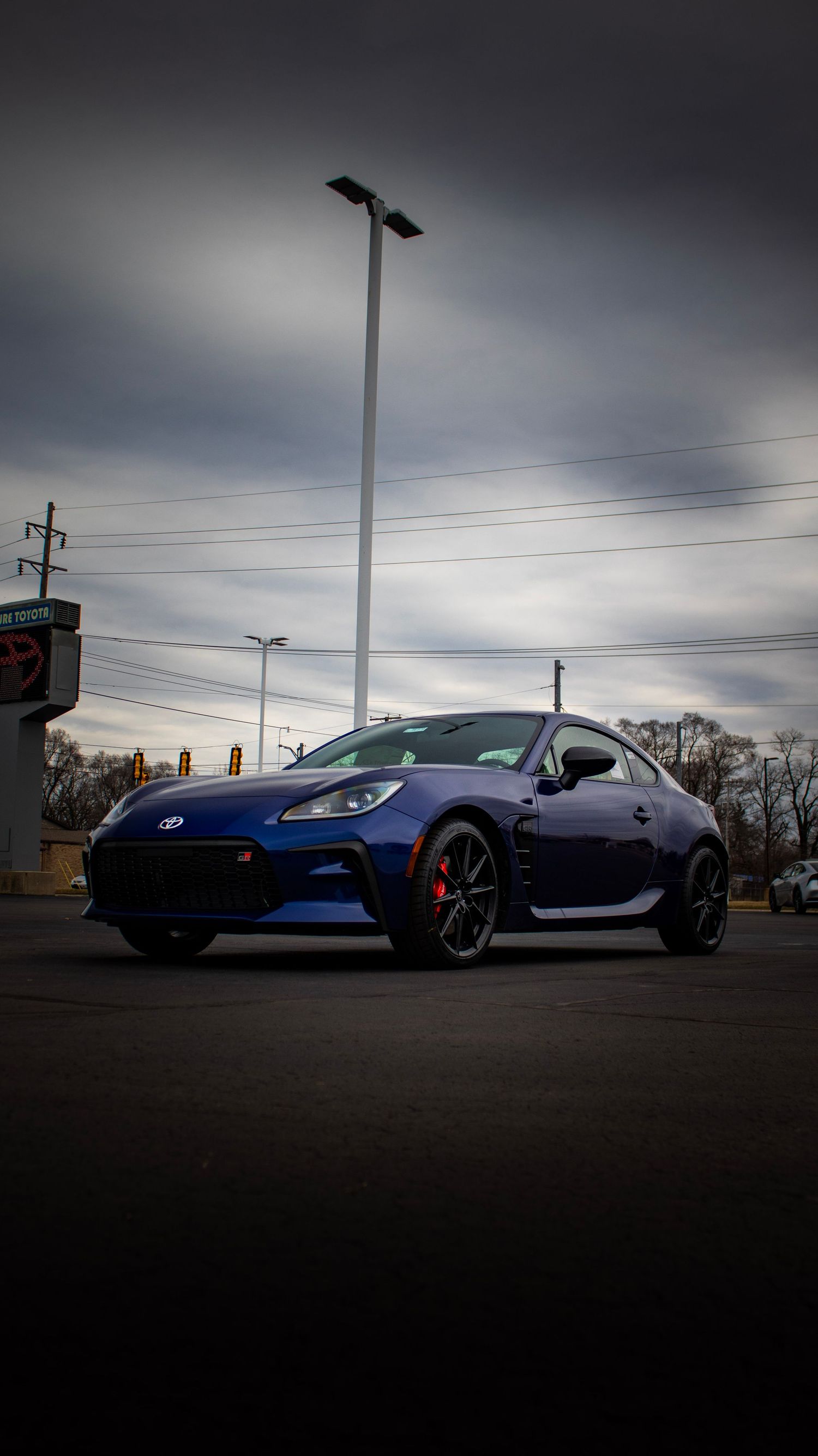 A blue Subaru BRZ sports car parked under a moody overcast sky with power lines and light poles in the background.