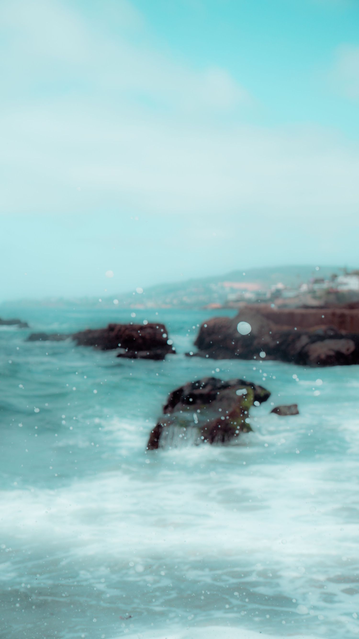 Moody seascape featuring dark rocks emerging from aqua waters under a pale cloudy sky.
