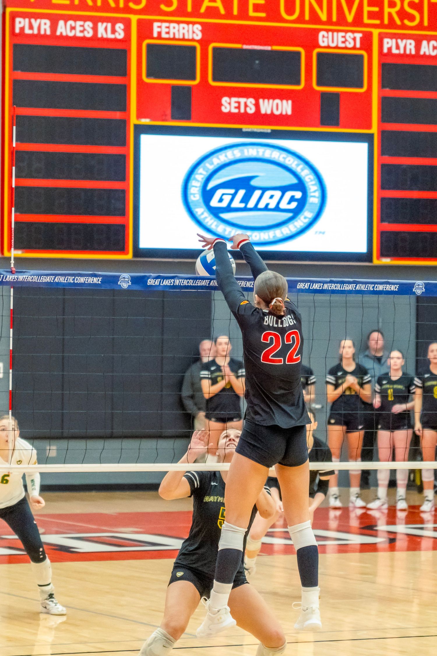 Volleyball players in black uniforms practice on a court with red scoreboard and university branding visible in background.