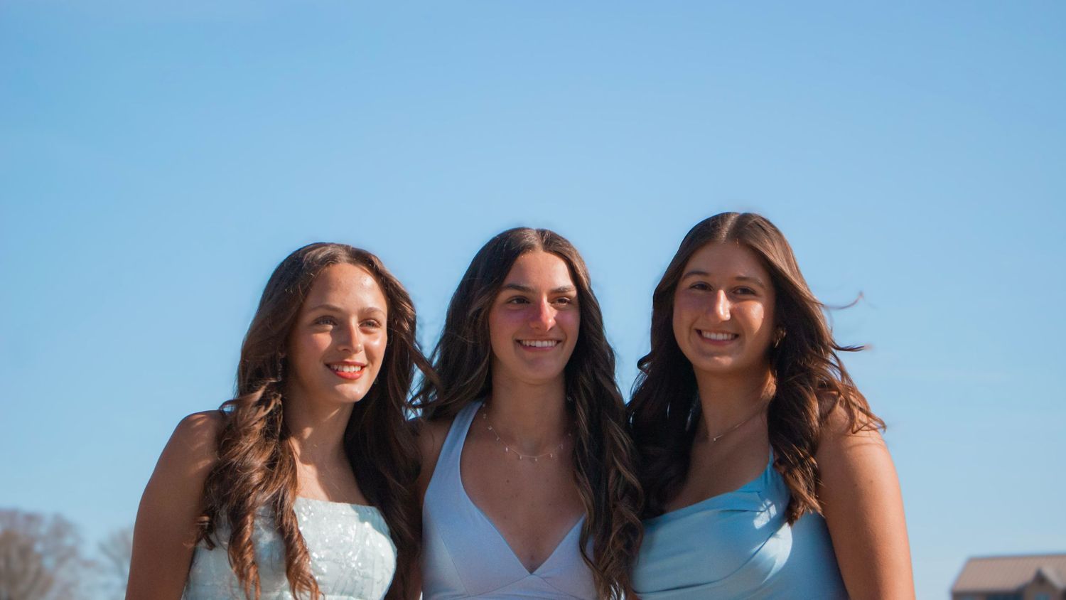Three friends in blue dresses pose together at a scenic beach overlook on a bright sunny day.