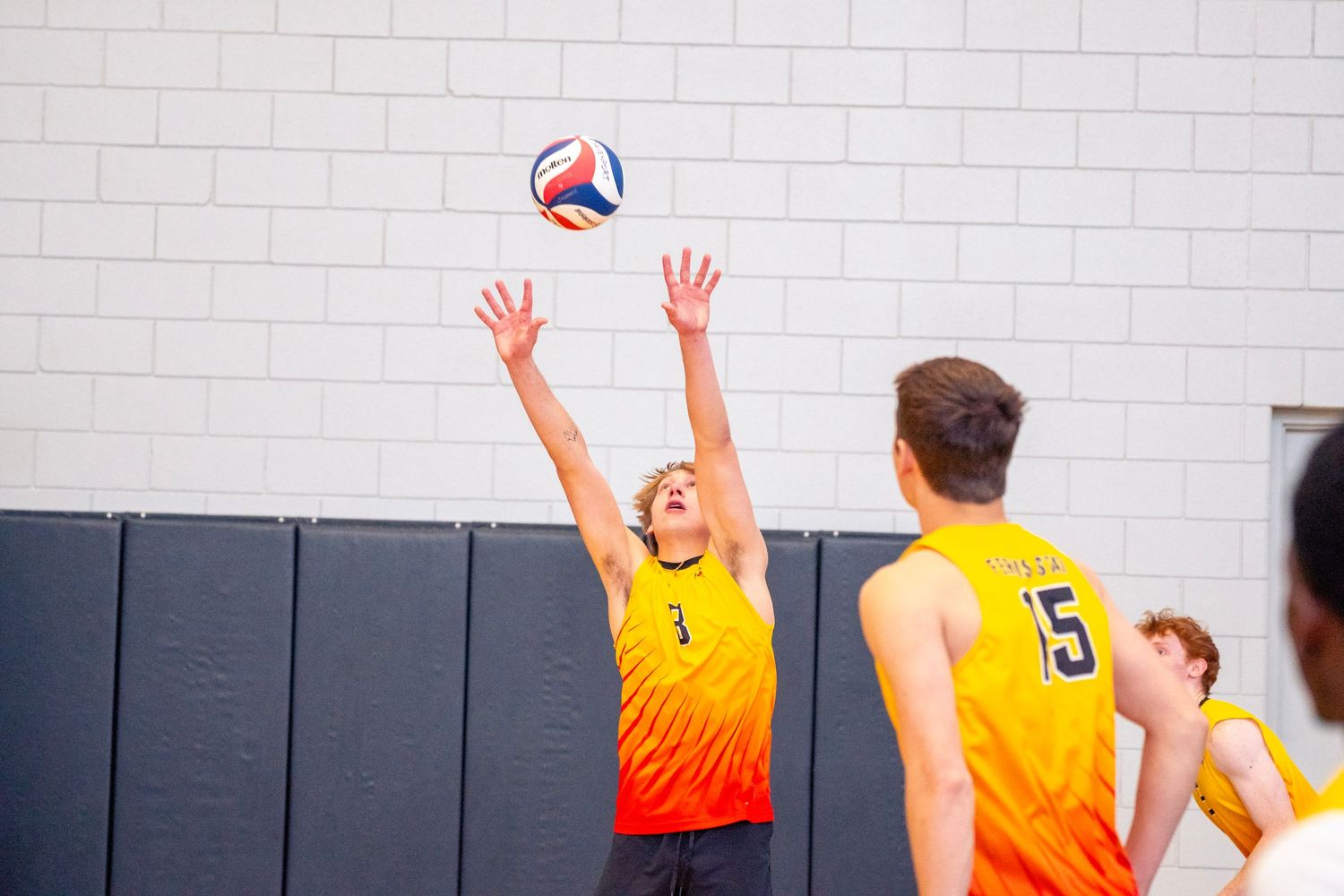 Volleyball players in bright orange jerseys compete at the net during an indoor match.