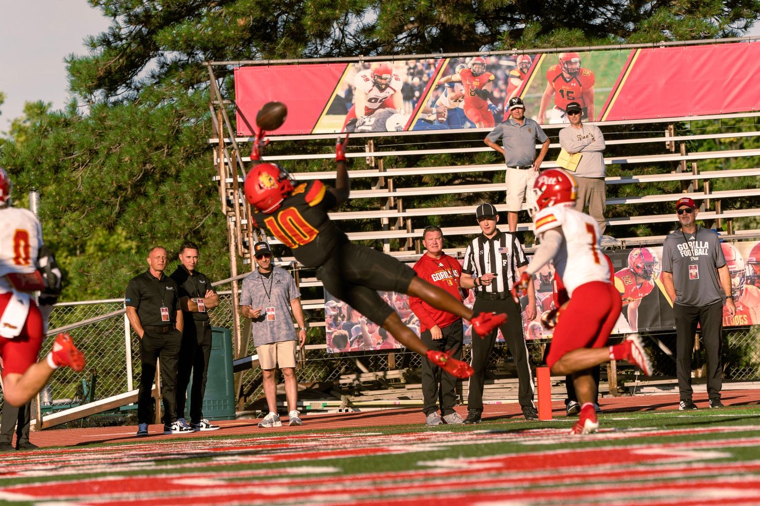 A panoramic view of a football field with red bleachers and spectators watching practice on a sunny day.