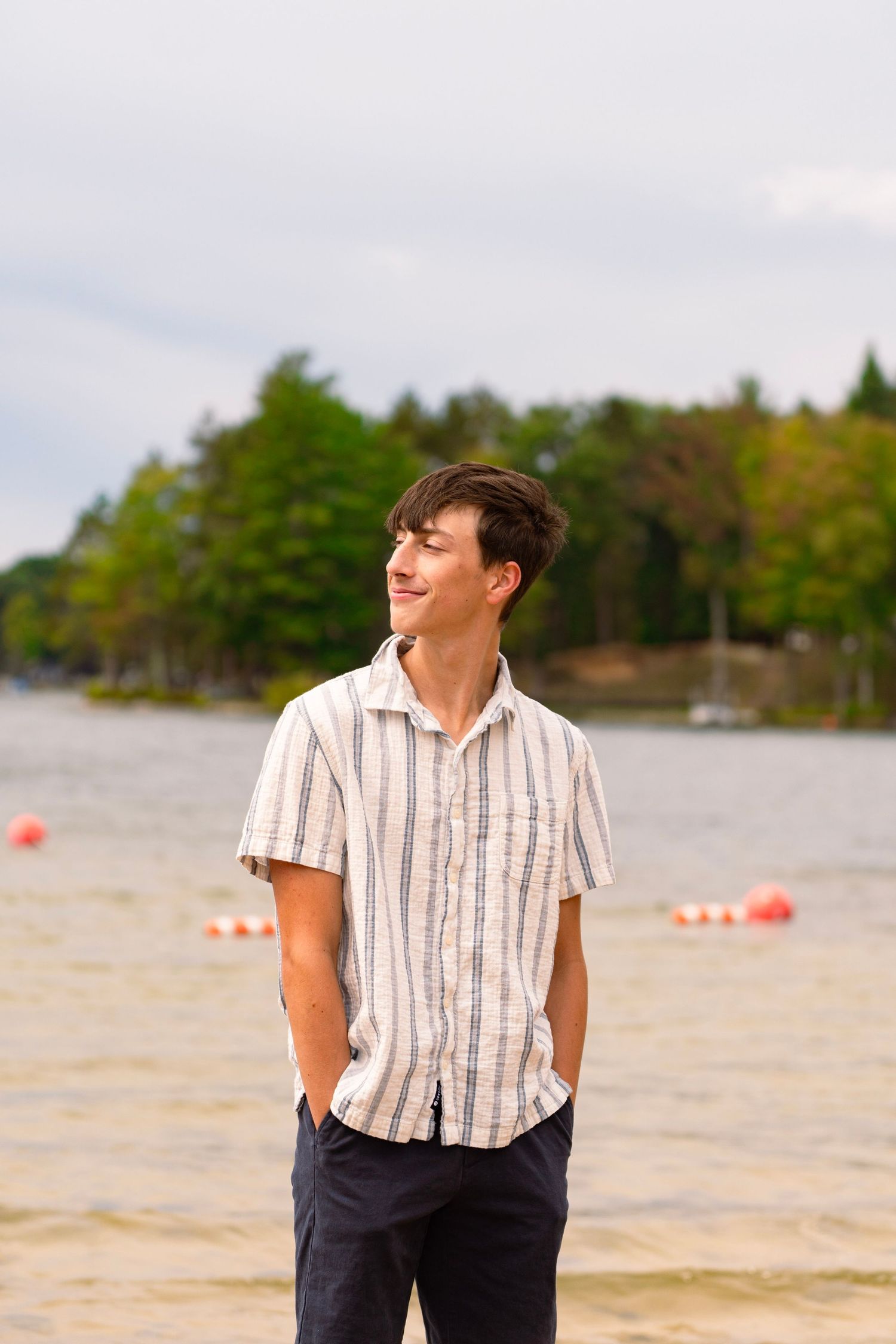 Smiling young man in a striped short-sleeve shirt stands with hands in pockets on a lakeside beach with buoys and trees behind him.