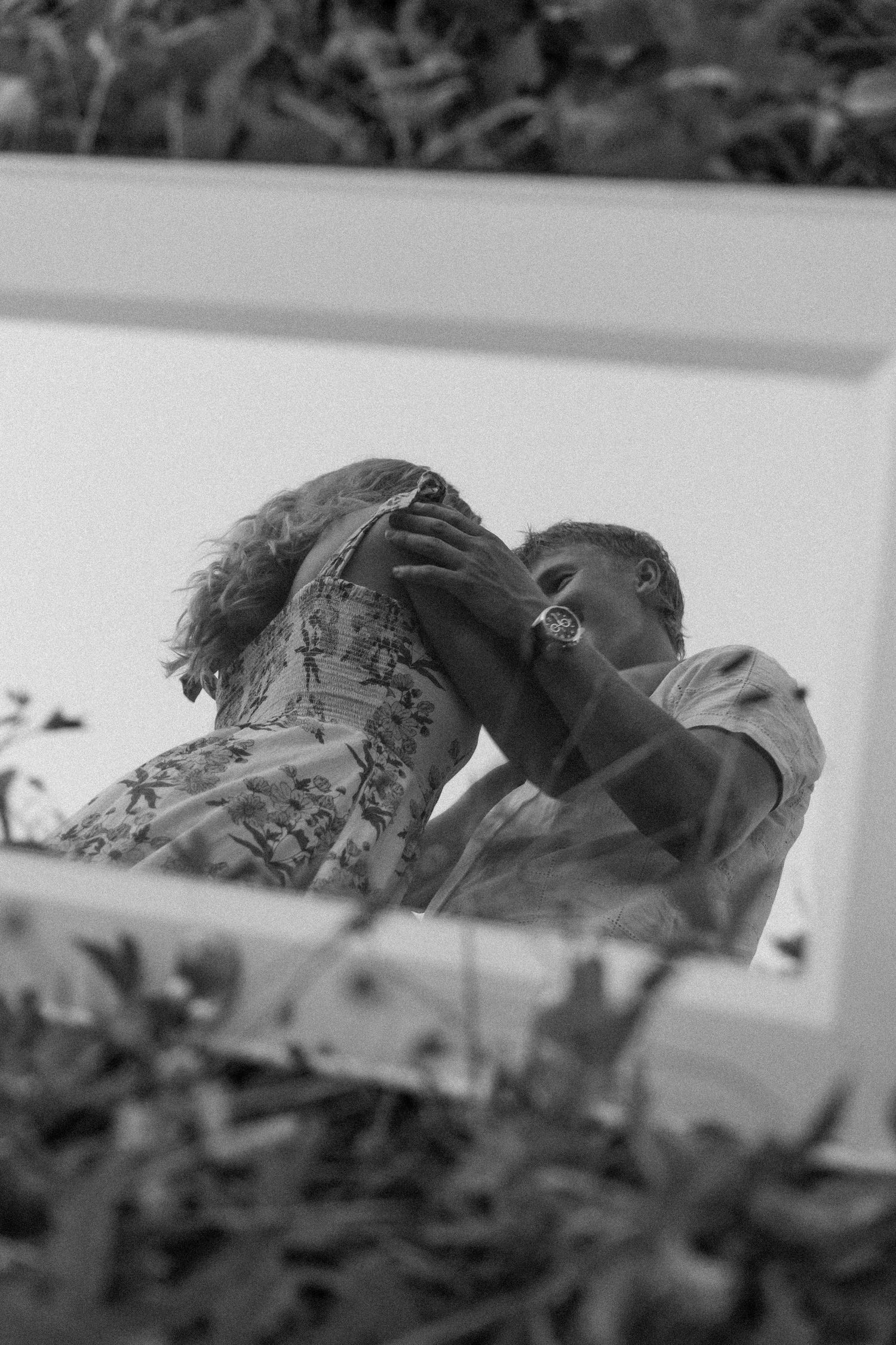 Black and white photo of a couple embracing, reflected in a mirror framed by leaves, woman in a floral dress and man wearing a watch.