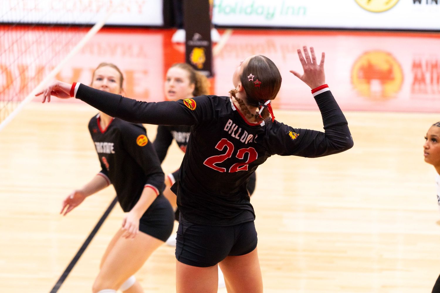Female volleyball player in a black number 22 jersey raises her arms mid-play near the net as teammates look on in an indoor gym.