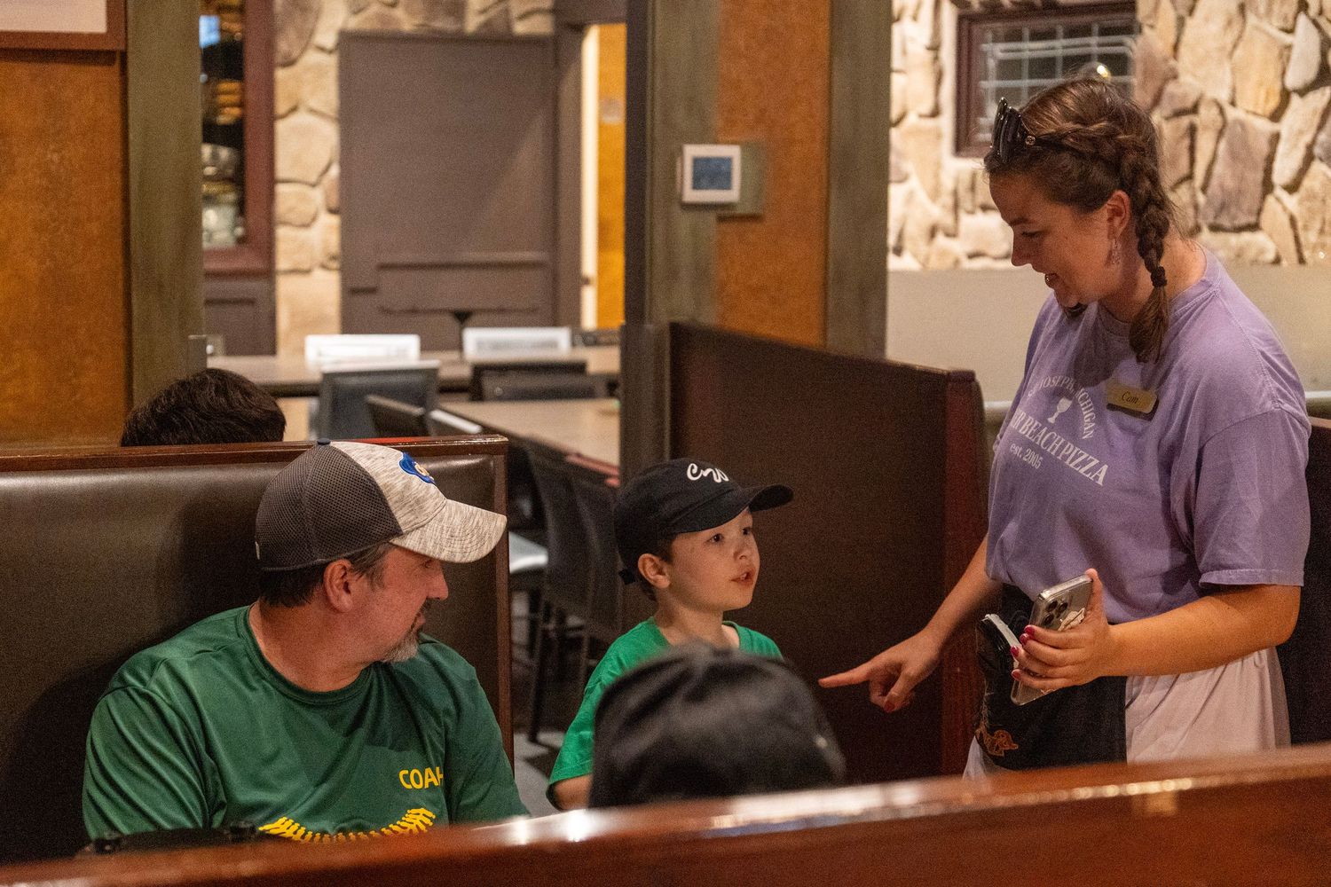 Staff in a purple shirt takes an order from customers seated at a restaurant booth with stone wall decor.