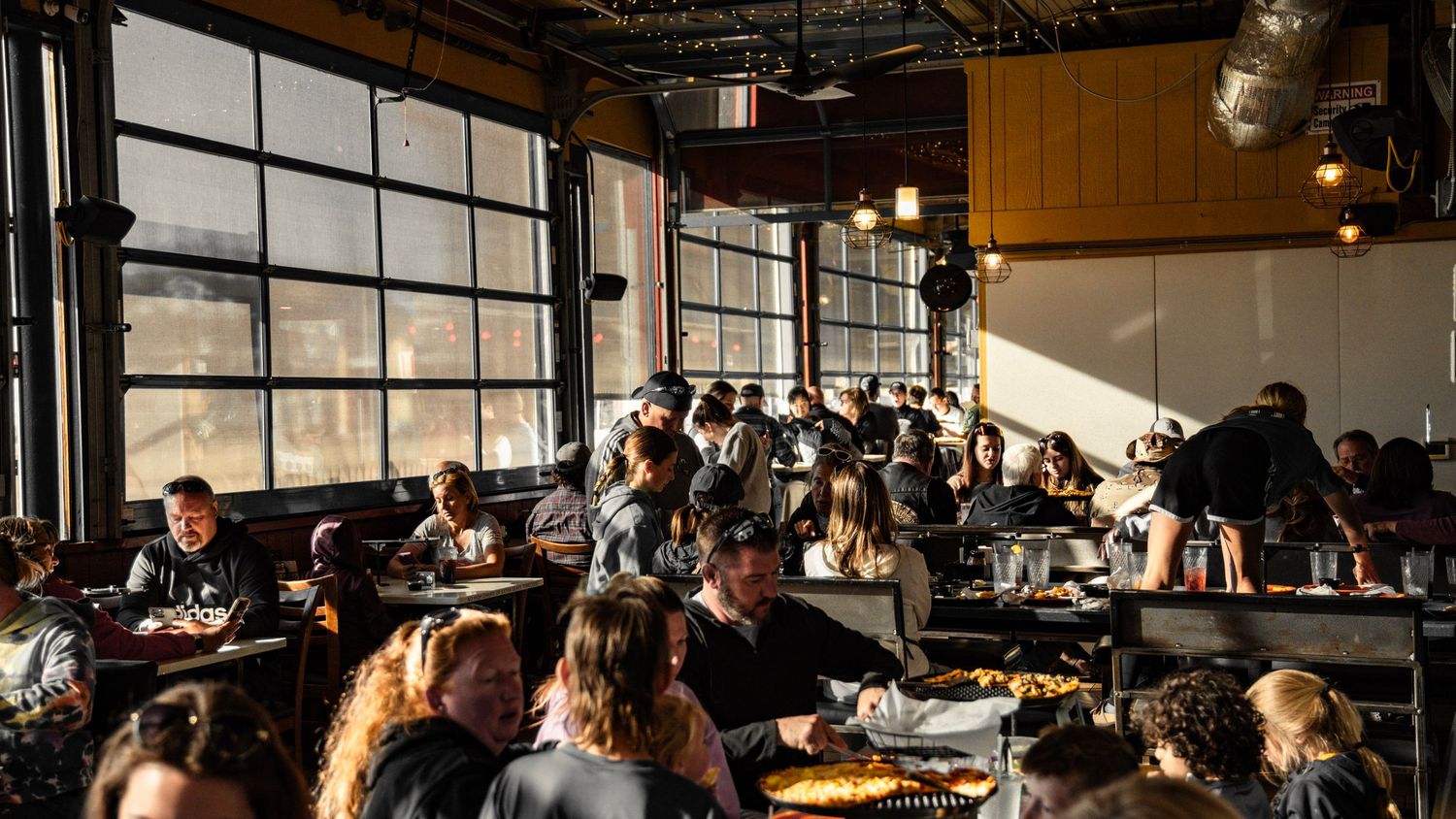 People working and studying in modern open space filled with natural light from large industrial-style windows.