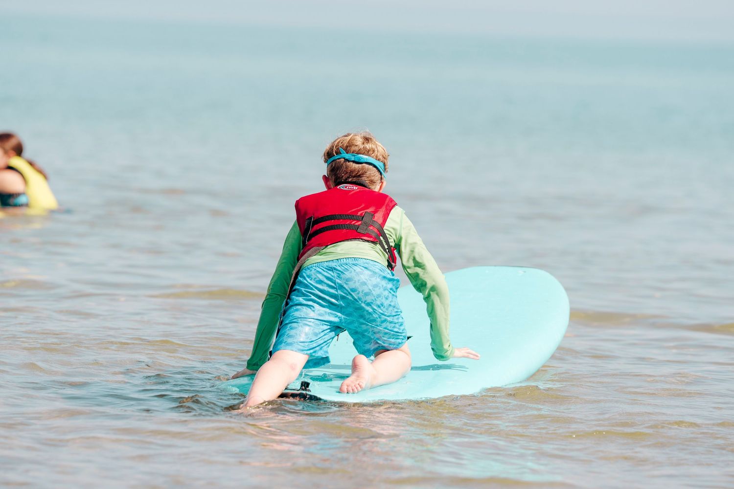 Child learning to surf lies on light blue surfboard in shallow water wearing red life vest.