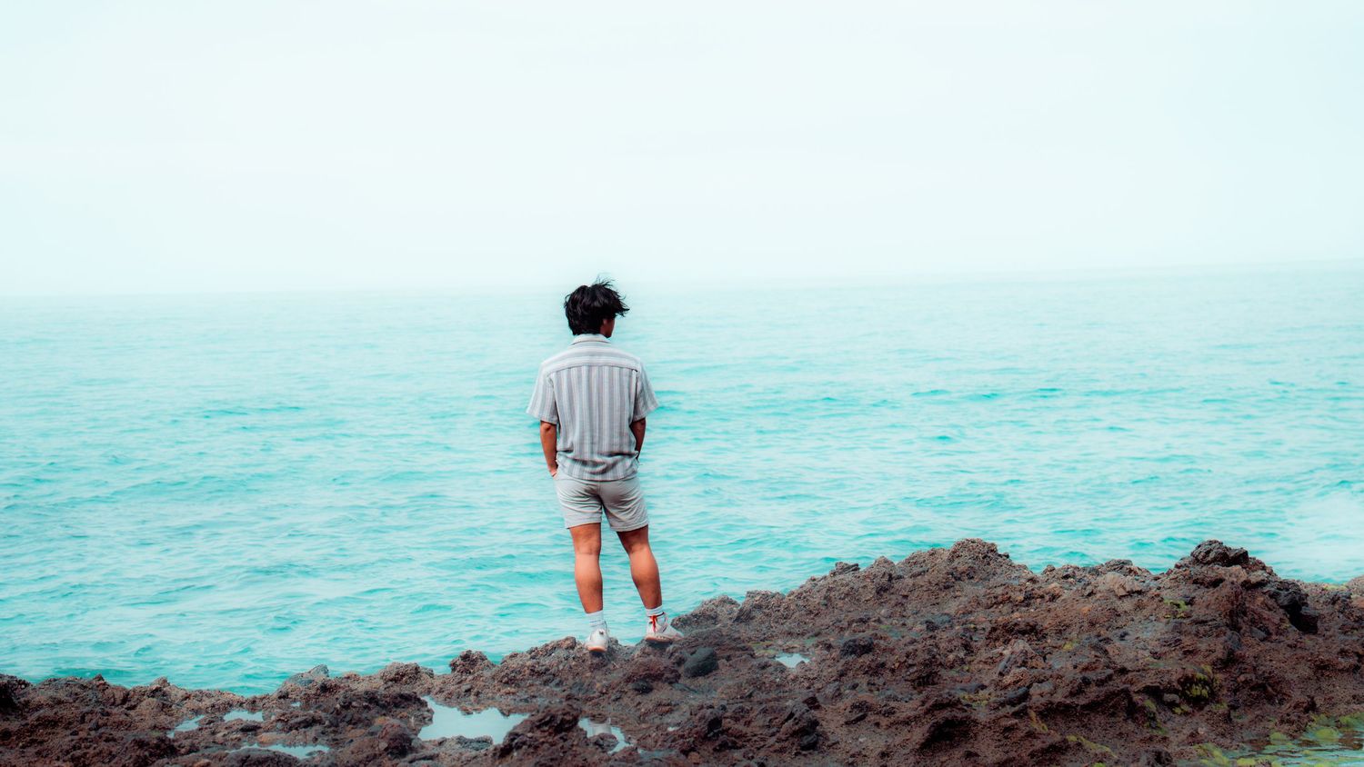 A figure stands alone on rocky coastal outcrop overlooking tranquil turquoise ocean waters on a hazy day.