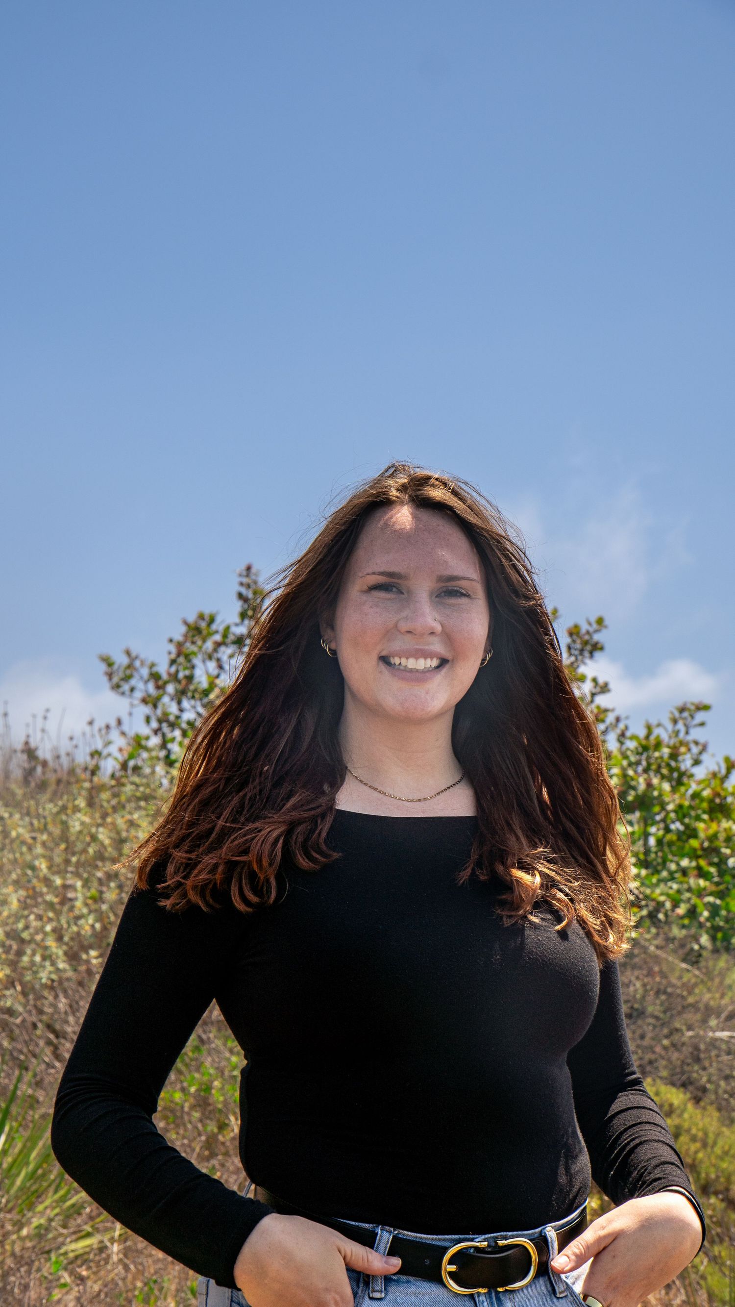 Person in black top and jeans smiling warmly while standing in outdoor natural setting.