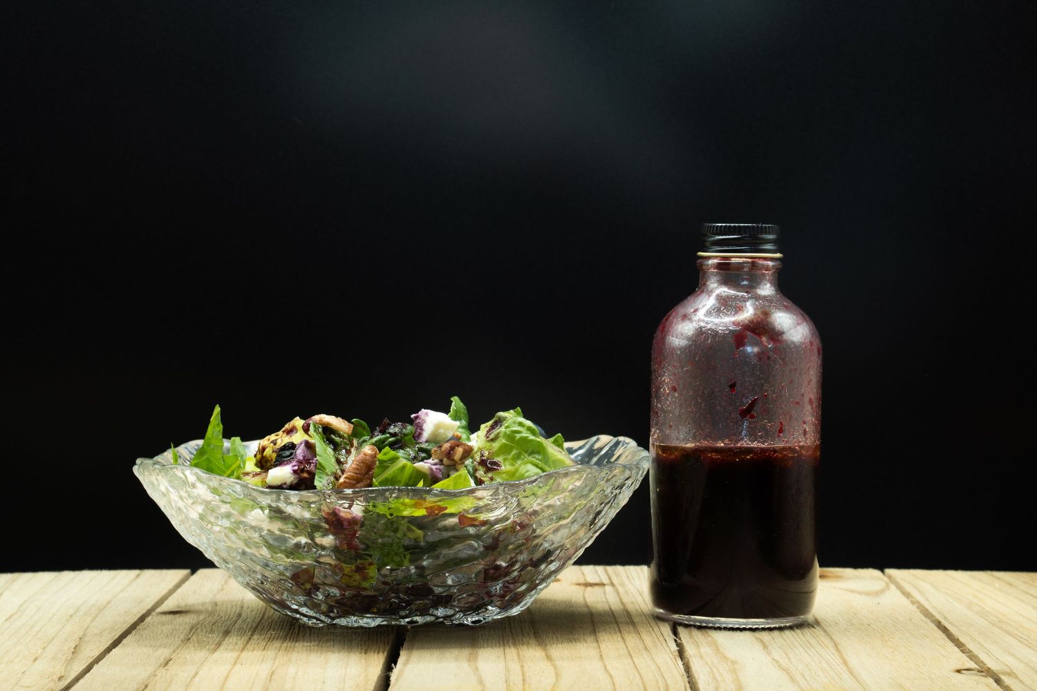 Crystal bowl of mixed green salad with dark vinaigrette dressing bottle on wooden table.