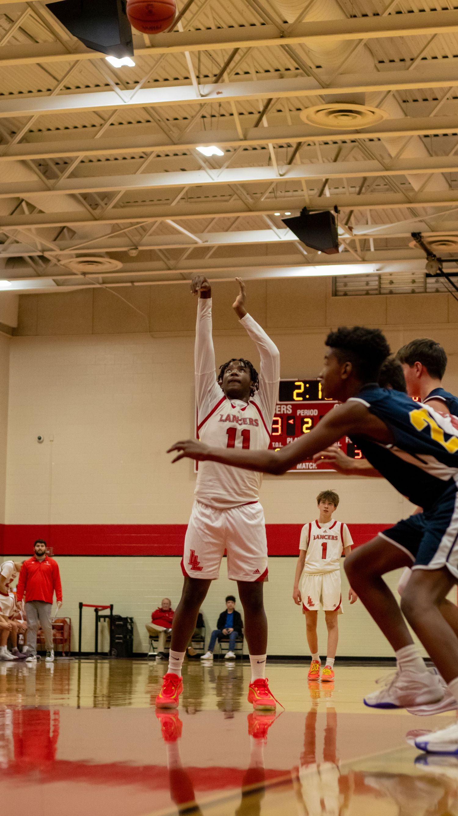 Basketball player in white jersey shoots while defender attempts to block.