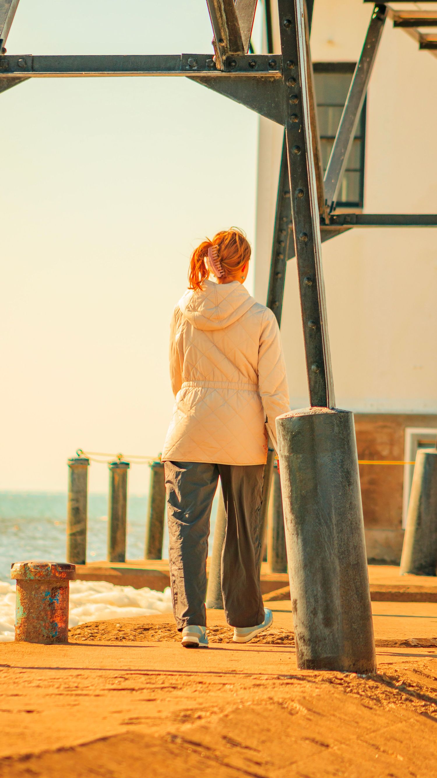 Person in beige jacket walking along pier with lighthouse in background.