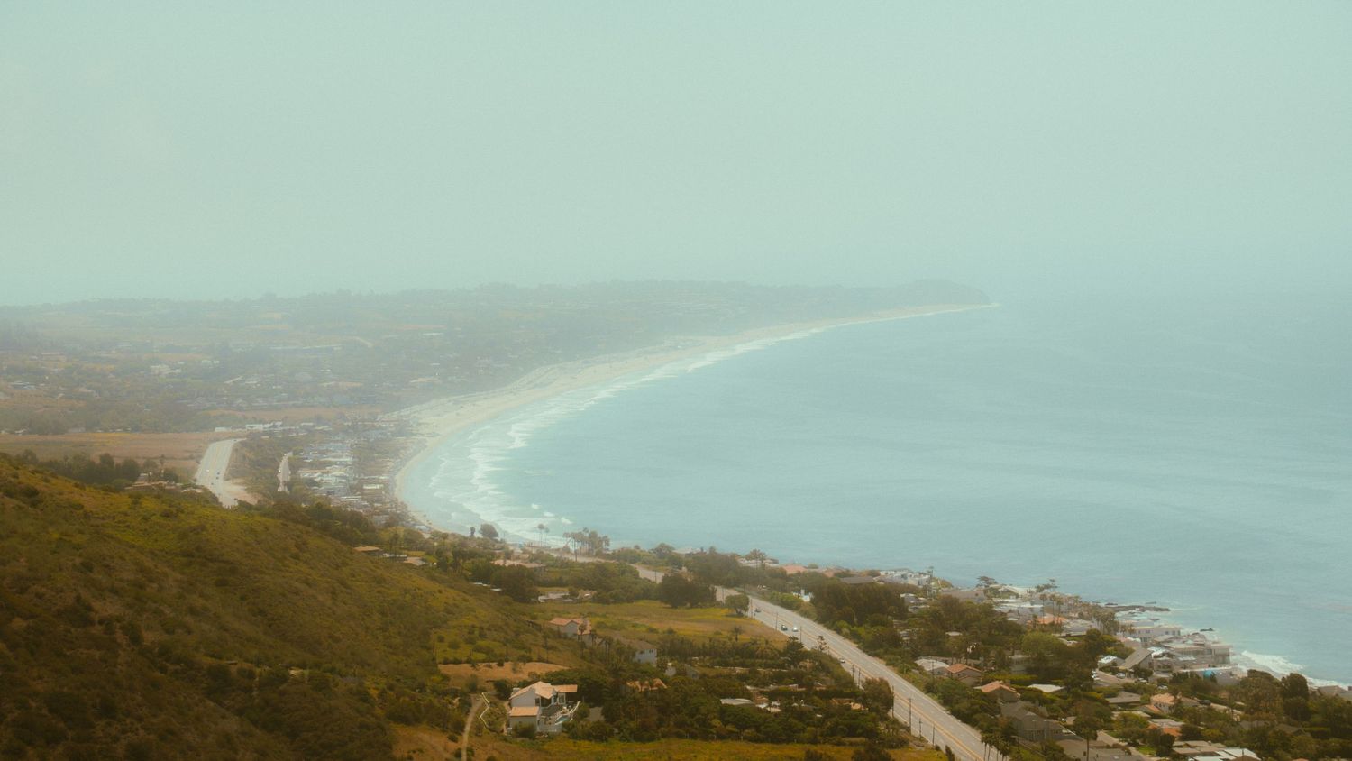 Misty coastal landscape with curved beach and residential buildings nestled in rolling hills.