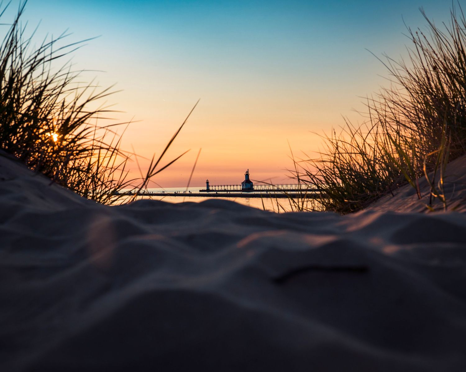 Silhouetted lighthouse and pier at sunset viewed through beach grass and sand dunes.