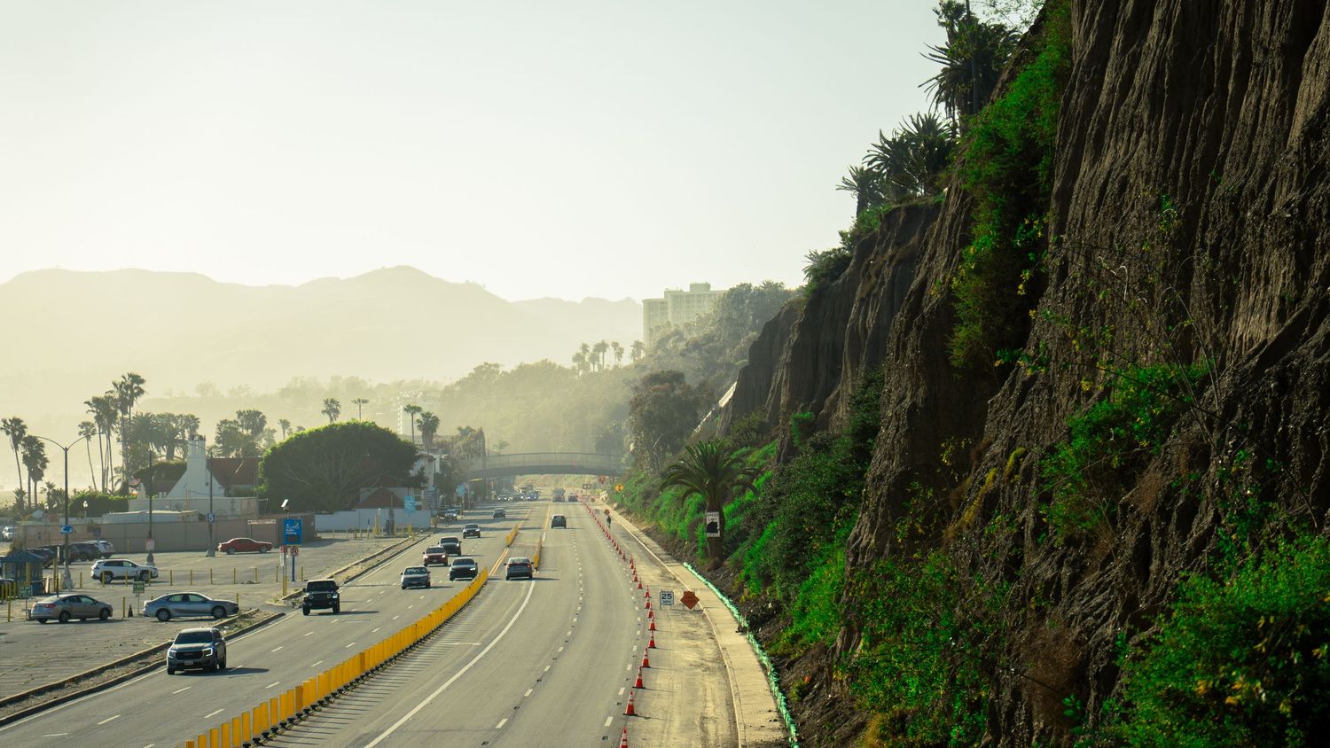Winding coastal road bordered by steep cliffs and vegetation with mountains in the distance.