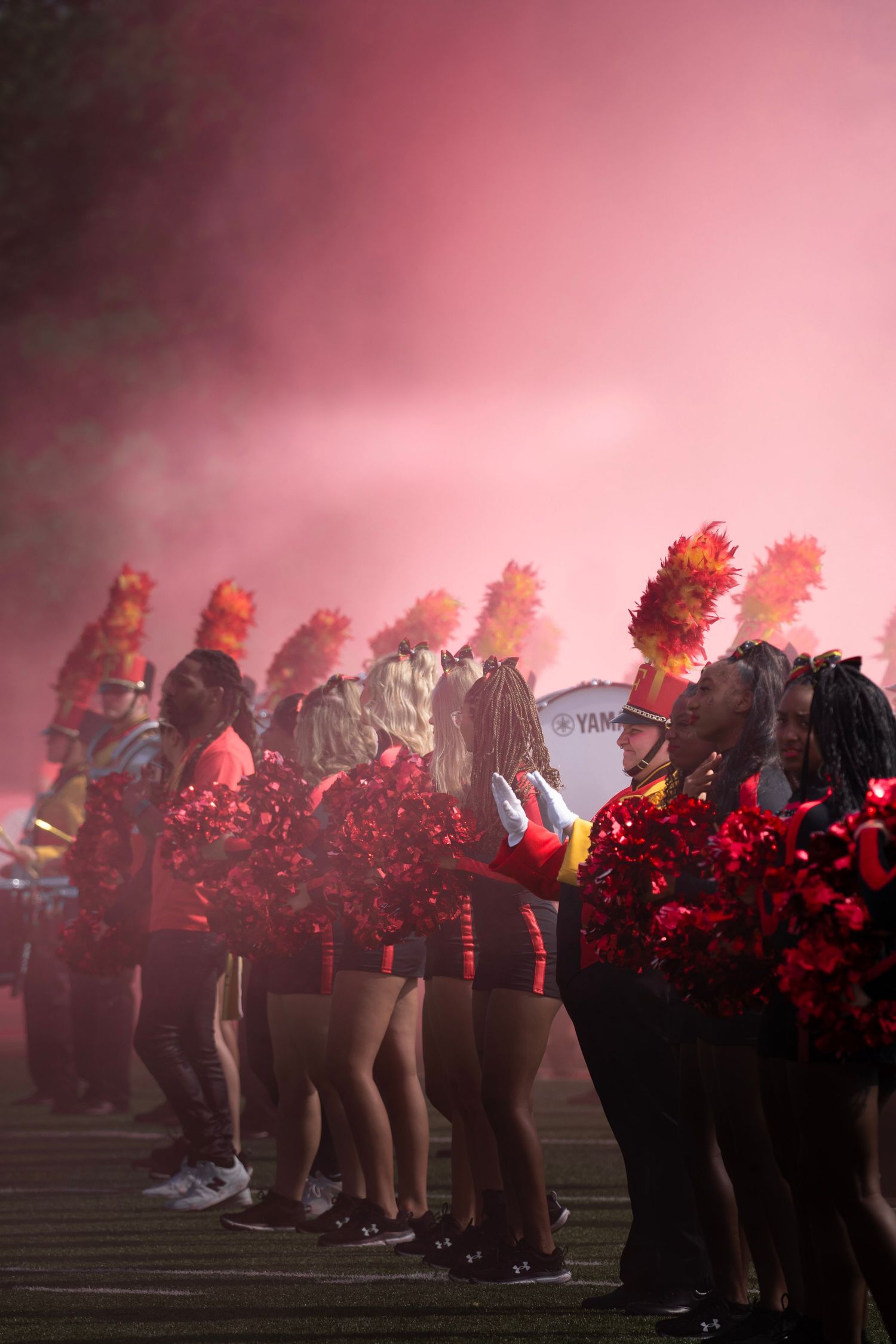 Cheerleaders perform in red smoke during a nighttime sporting event with pom poms and dramatic lighting effects.