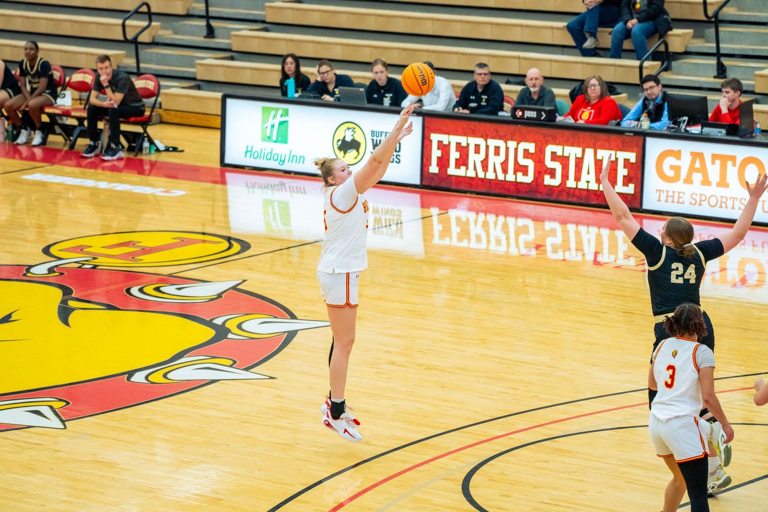 Basketball player in white jersey takes a jump shot during game at Ferris State University gymnasium.