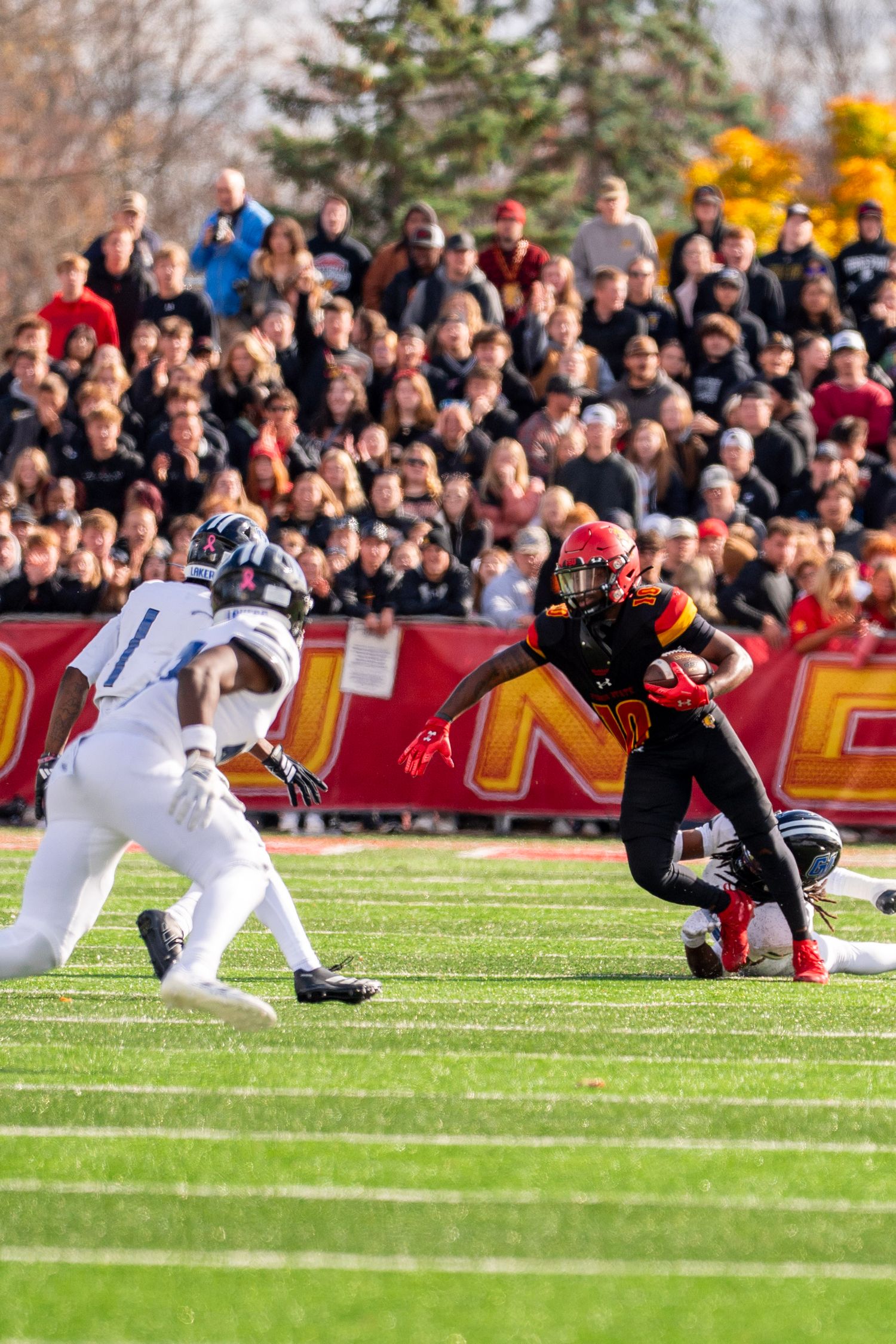 Football players collide during a tackle sequence in front of a large crowd of spectators on a sunny autumn day.