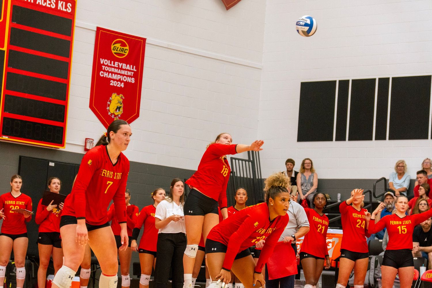 A volleyball team in red uniforms celebrates together on a court with red banners and spectators in the background.