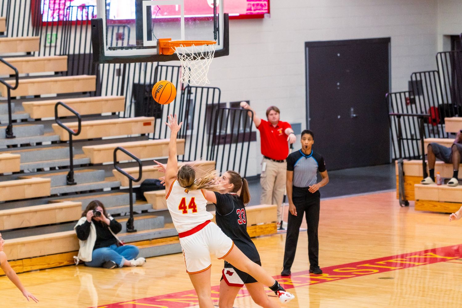 Basketball players compete during an indoor game with spectators watching from wooden bleachers in a gymnasium.