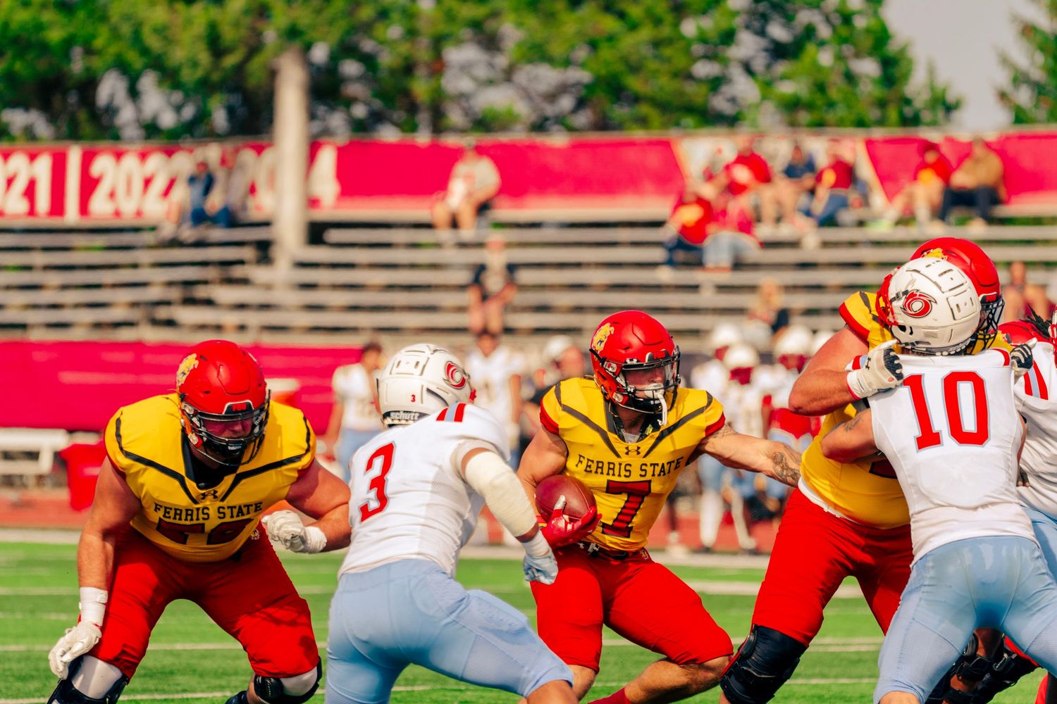 Football players in yellow and red uniforms engage in an intense play during a game at a stadium with bleachers visible.