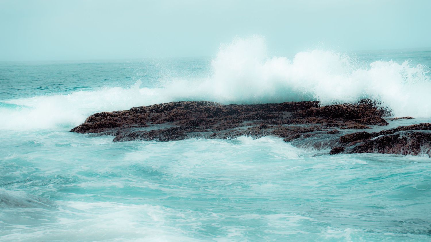 Dramatic wave spray erupts as turquoise waters collide with a dark rocky outcrop in the ocean.