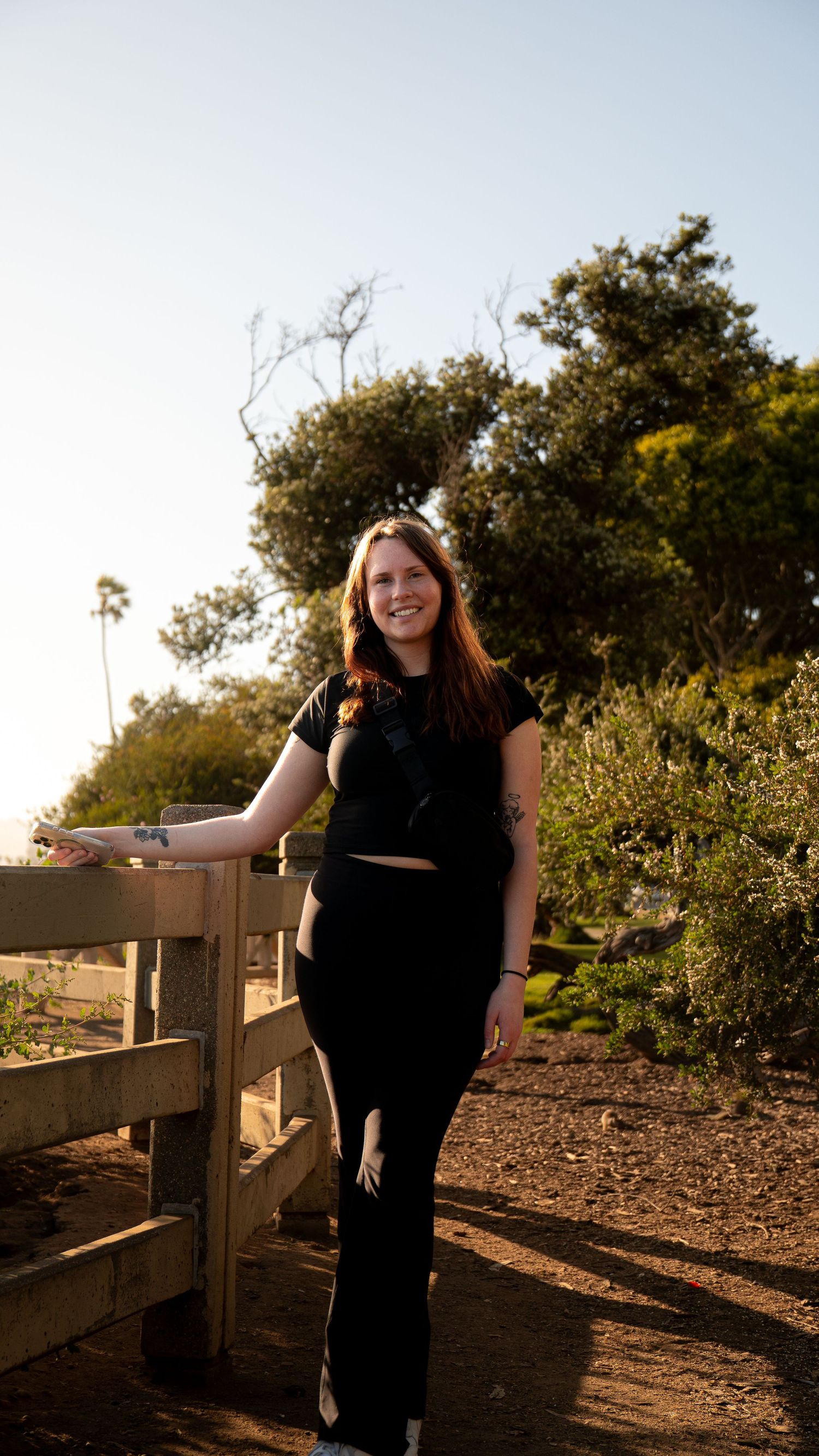 Person in black outfit poses along rustic wooden fence during sunset.