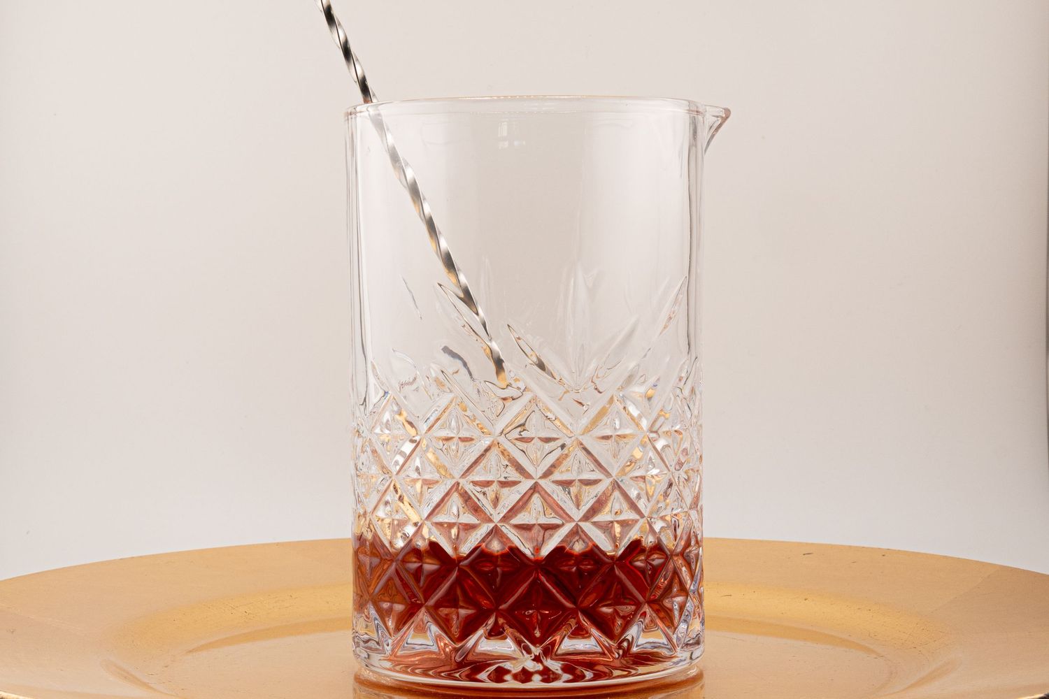 Crystal mixing glass with red syrup and black striped straw on gold plate.