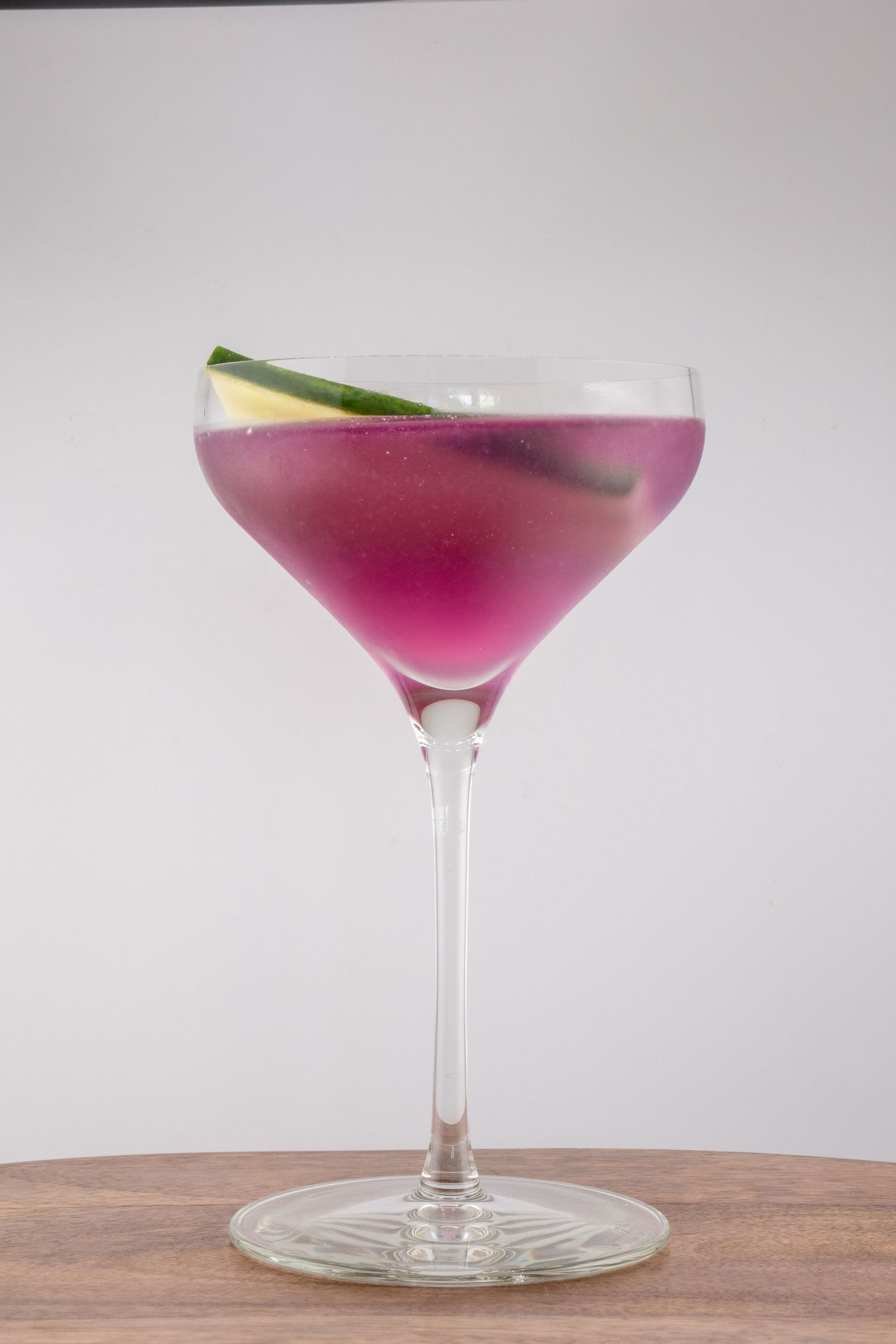 Vibrant magenta cocktail in stemmed glass with cucumber garnish on wooden table.