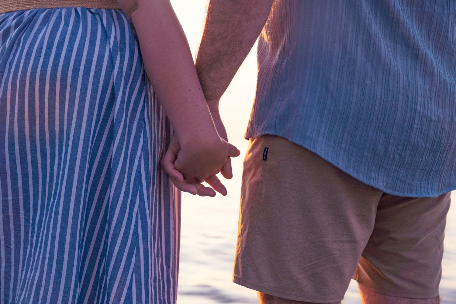 Two people holding hands at sunset, wearing a striped dress and shorts on a beach.